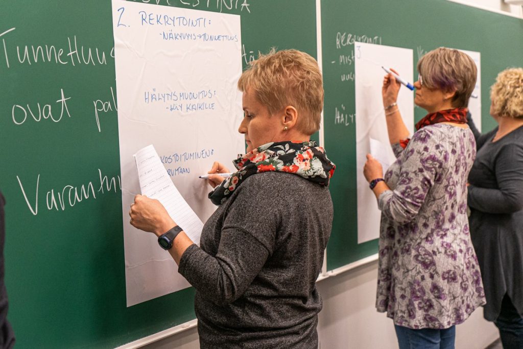 Kuvassa kolme työpajaan osallistunutta kirjoittavat ideoitaan ylös liitutaululle. 