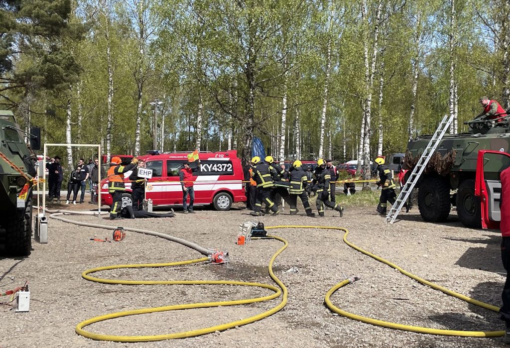 Kuvassa Jehumalja-kilpailun kalusto-osio on käynnissä. Kuvien reunoilla näkyy armeijan kalustoa, keskellä on ambulanssi. Pelastaja-asuiset kilpailijat kantavat paaria juosten kohti ambulanssia. Kuva: Länsi-Uudenmaan pelastuslaitos