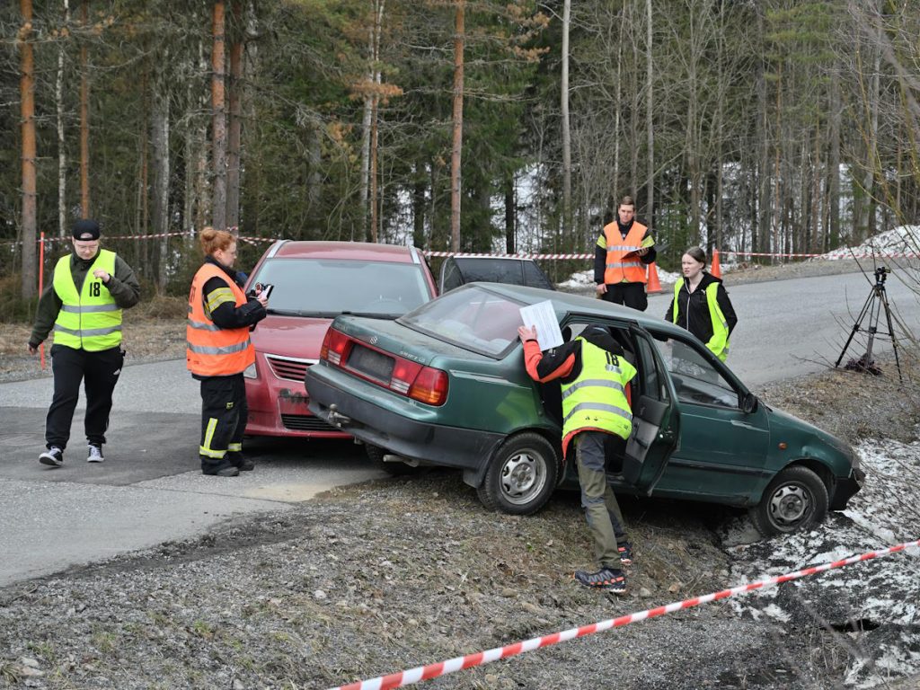 Kuva NouHätä! 2023 -kilpailun finaalin onnettomuusrastilta, jossa punainen auto on törmännyt vihreän auton takakylkeen. Kuvassa kolme keltaliivistä kilpailijaa selvittää tilannetta ja kaksi oranssiliivistä fasilitoijaa tarkkailee kilpailutilanteen etenemistä. Kuva: SPEK ry. 