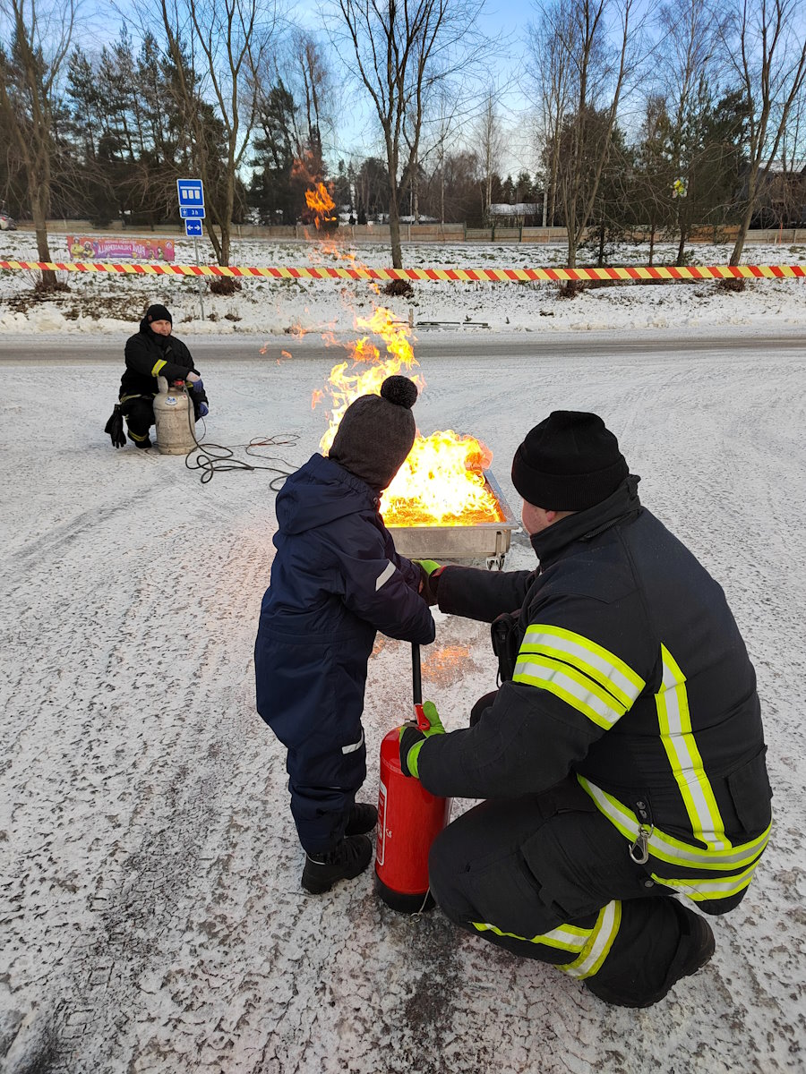 Kuvassa lapsi sammuttaa parkkipaikalla vaahtosammuttimella pelastajan avustamana laatikossa olevaa paloa. Kuva: SPEK ry