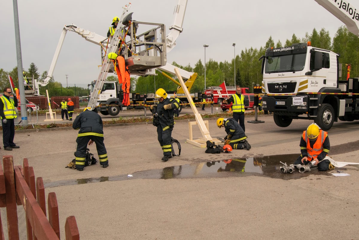 Kuva kilpailutilanteesta, jossa on paljon meneillään. Yksi kilpailija on paloauton nostokorissa, toinen asettelee tikkaita nostokoria vasten. Yksi kilpailija sahaa puutavaraa ja yksi asentaa letkusuutinta.