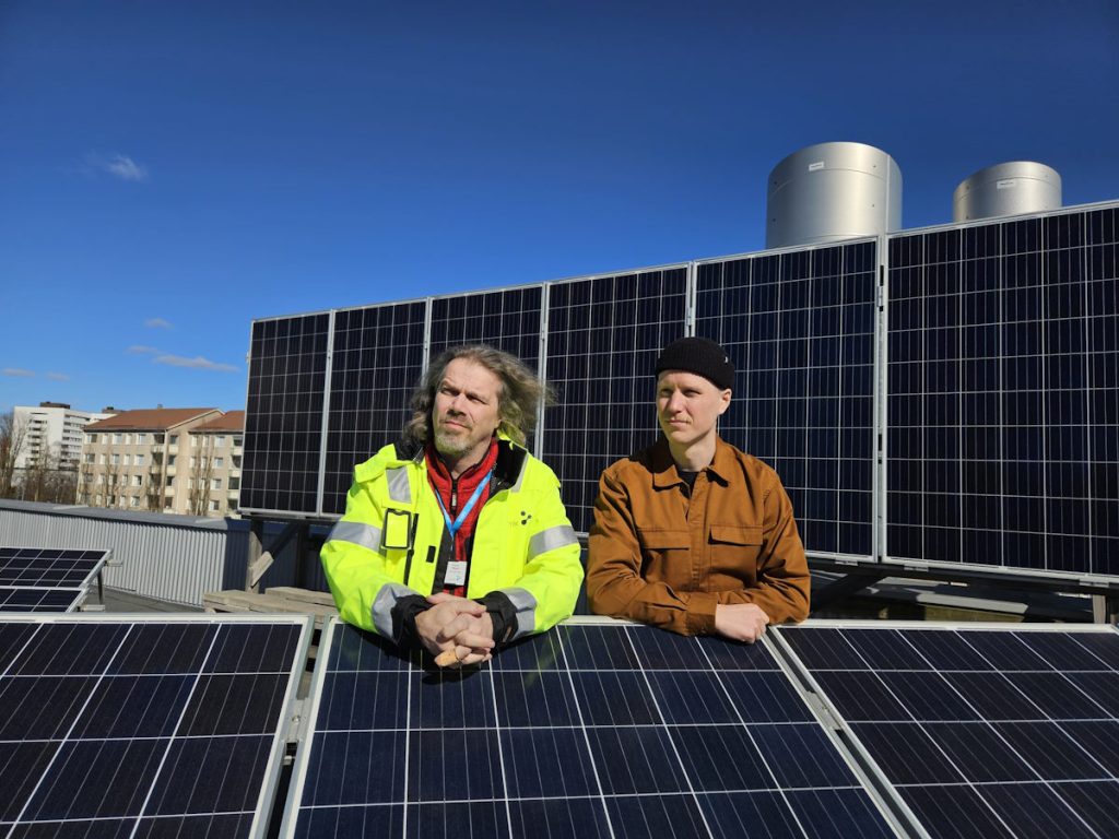 Kuvan henkilöt poseeraavat kameralle aurinkoisena päivänä talon katolla aurinkopaneelien keskellä. 